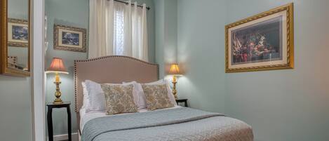 A bedroom with a bed, two bedside tables with lamps, a window with curtains, and framed pictures on the walls.