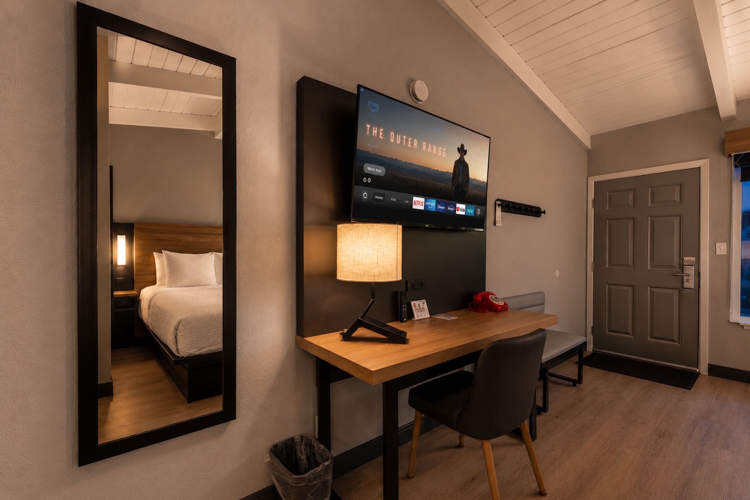 Une chambre d’hôtel avec un lit, un bureau doté d’une lampe, un téléviseur diffusant la série « The Outer Range », et une porte.