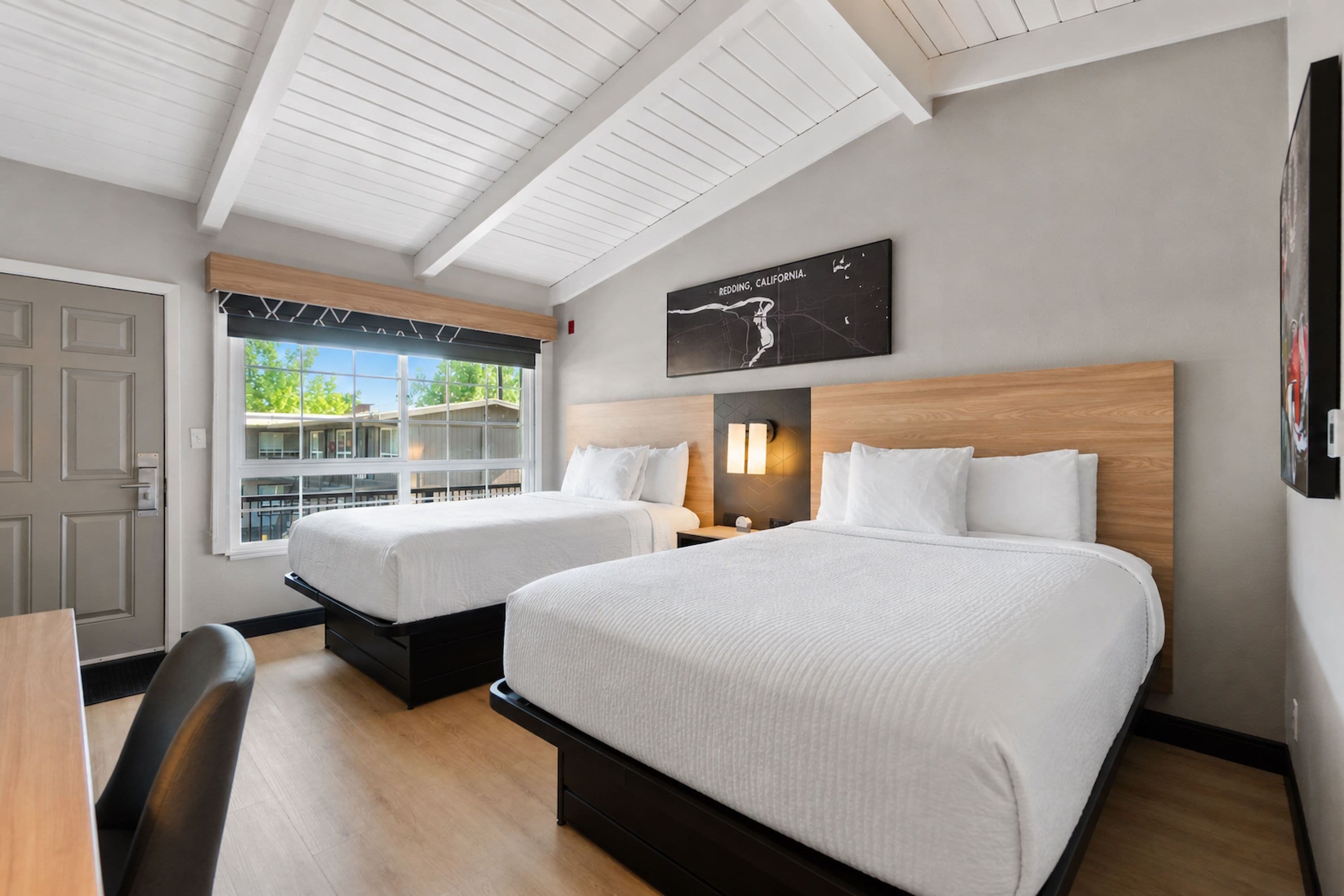 A hotel room with two beds, a desk, and a window with a view of a building.