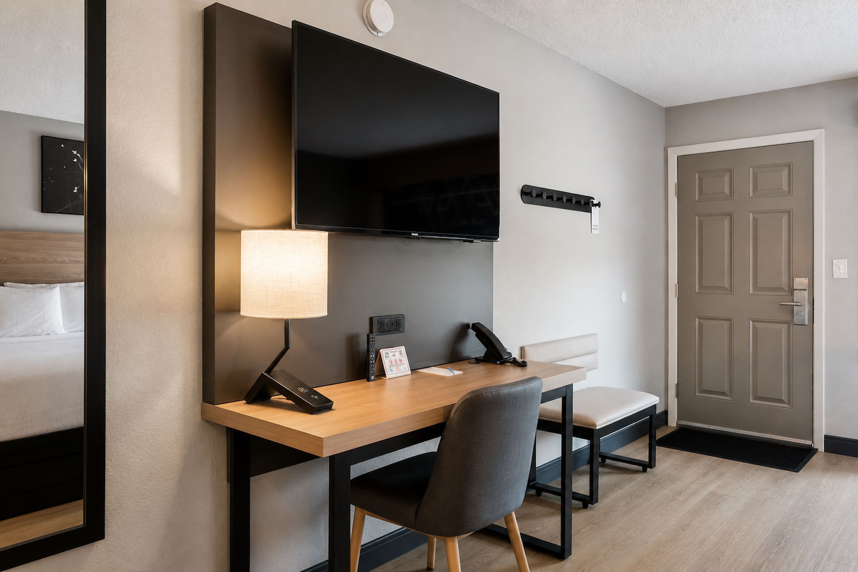 A hotel room with a bed, a desk with a chair, a flat-screen TV, a lamp, a telephone, and a door.