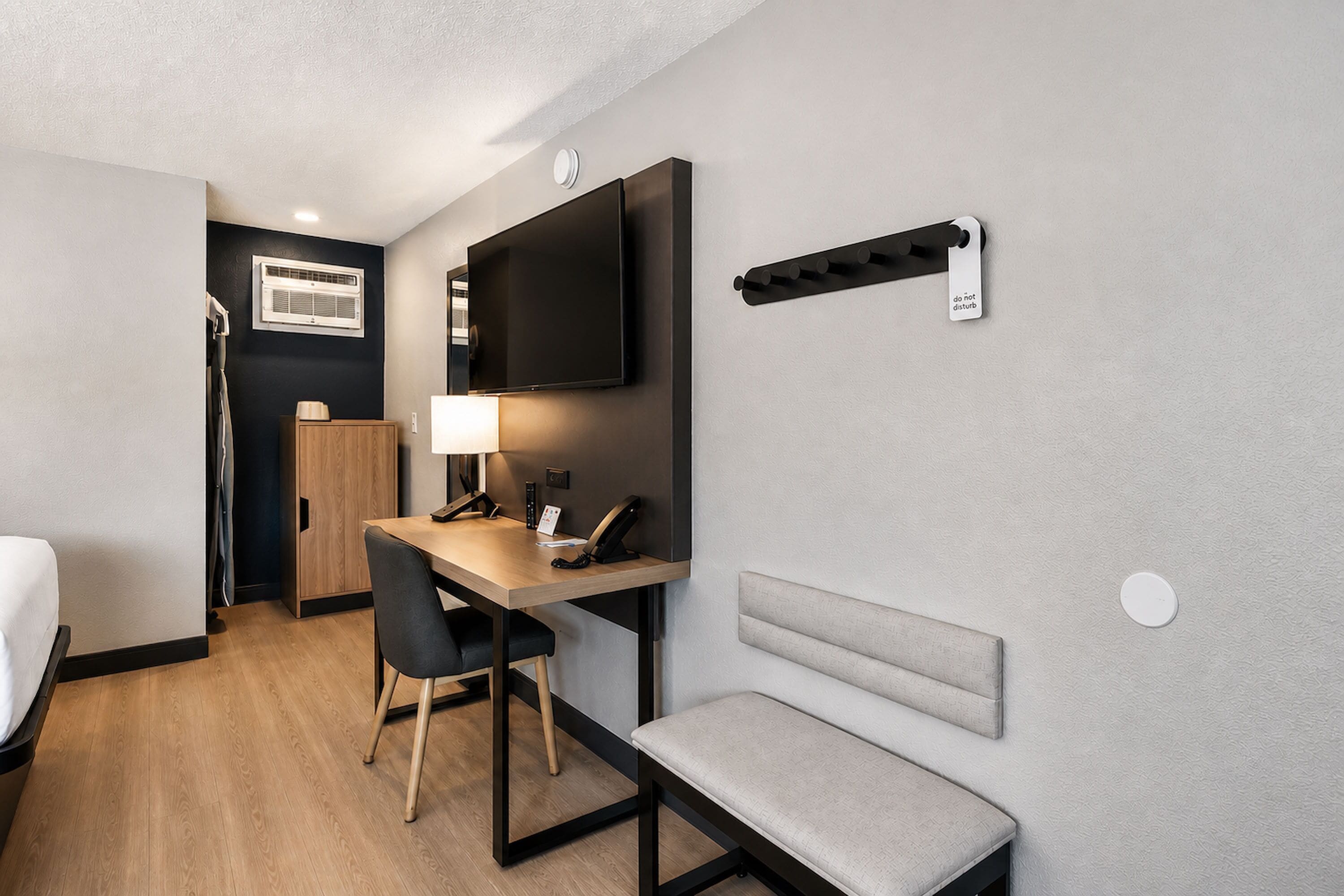 A hotel room with a bed, a desk with a chair, a flat-screen TV, a bedside table, and a wall-mounted shelf.