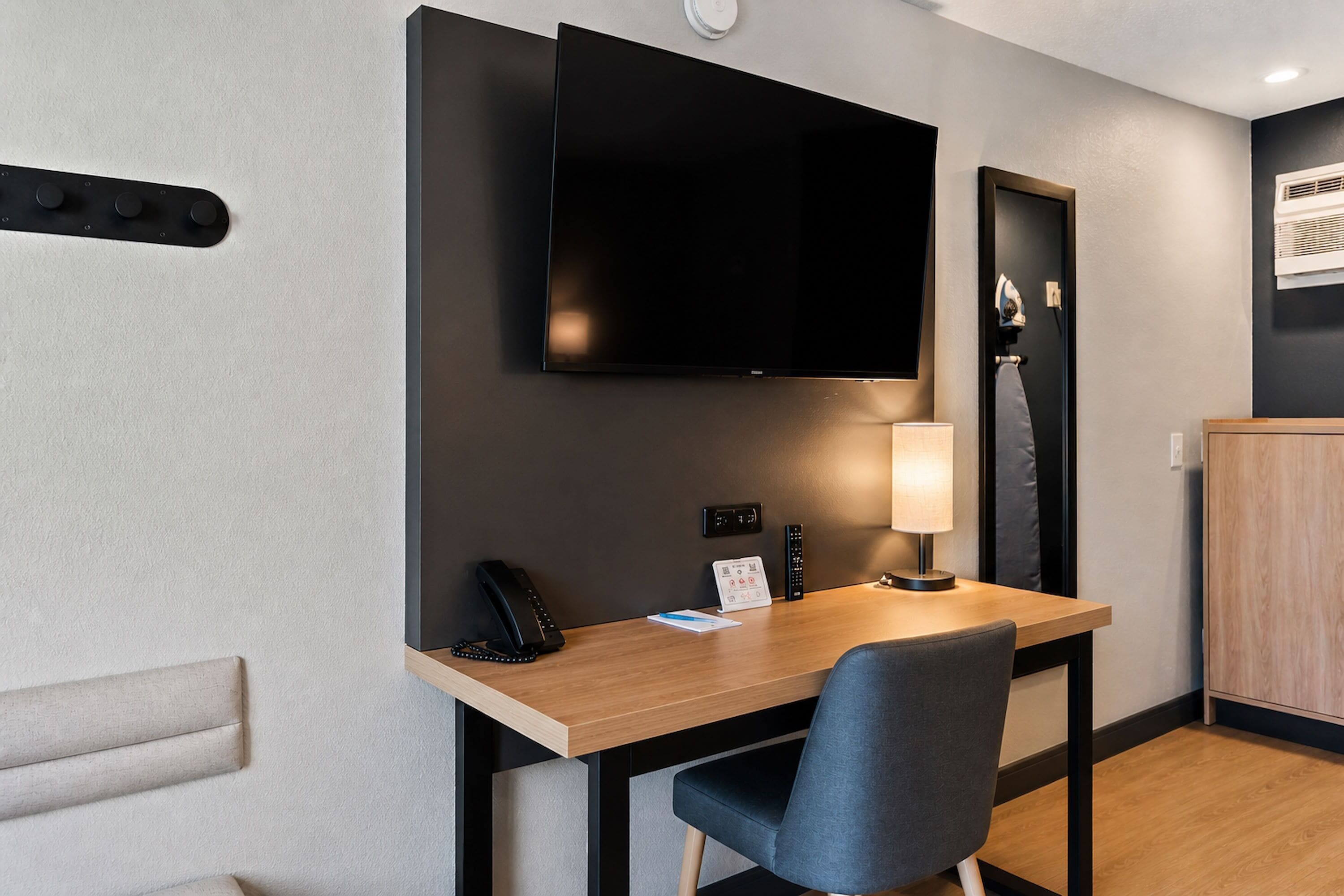 A hotel room with a desk, chair, flat-screen TV, bedside table, lamp, and a wardrobe.