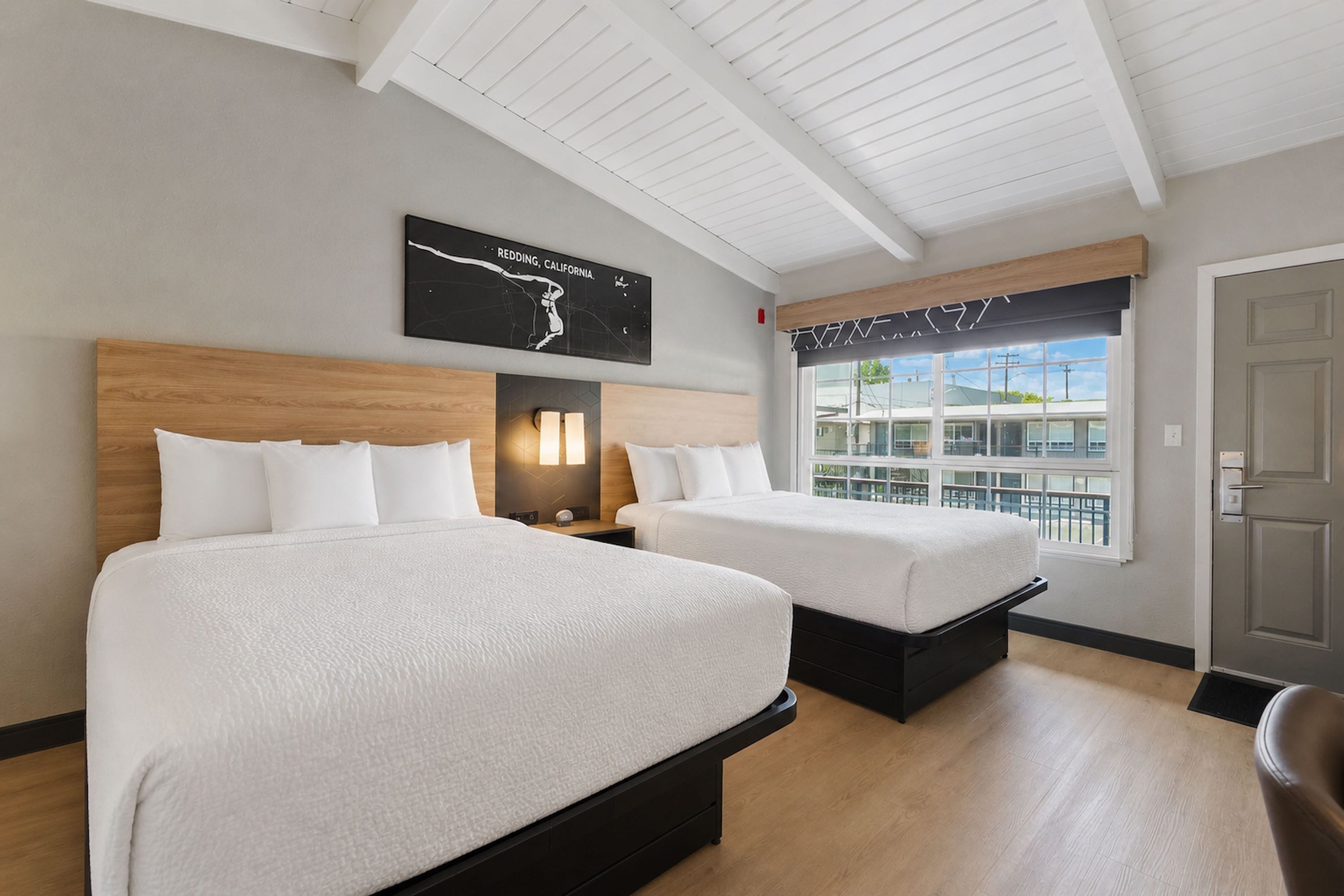 A hotel room with two beds, a wooden headboard, a nightstand with a lamp, a window with a view of buildings, and a door.