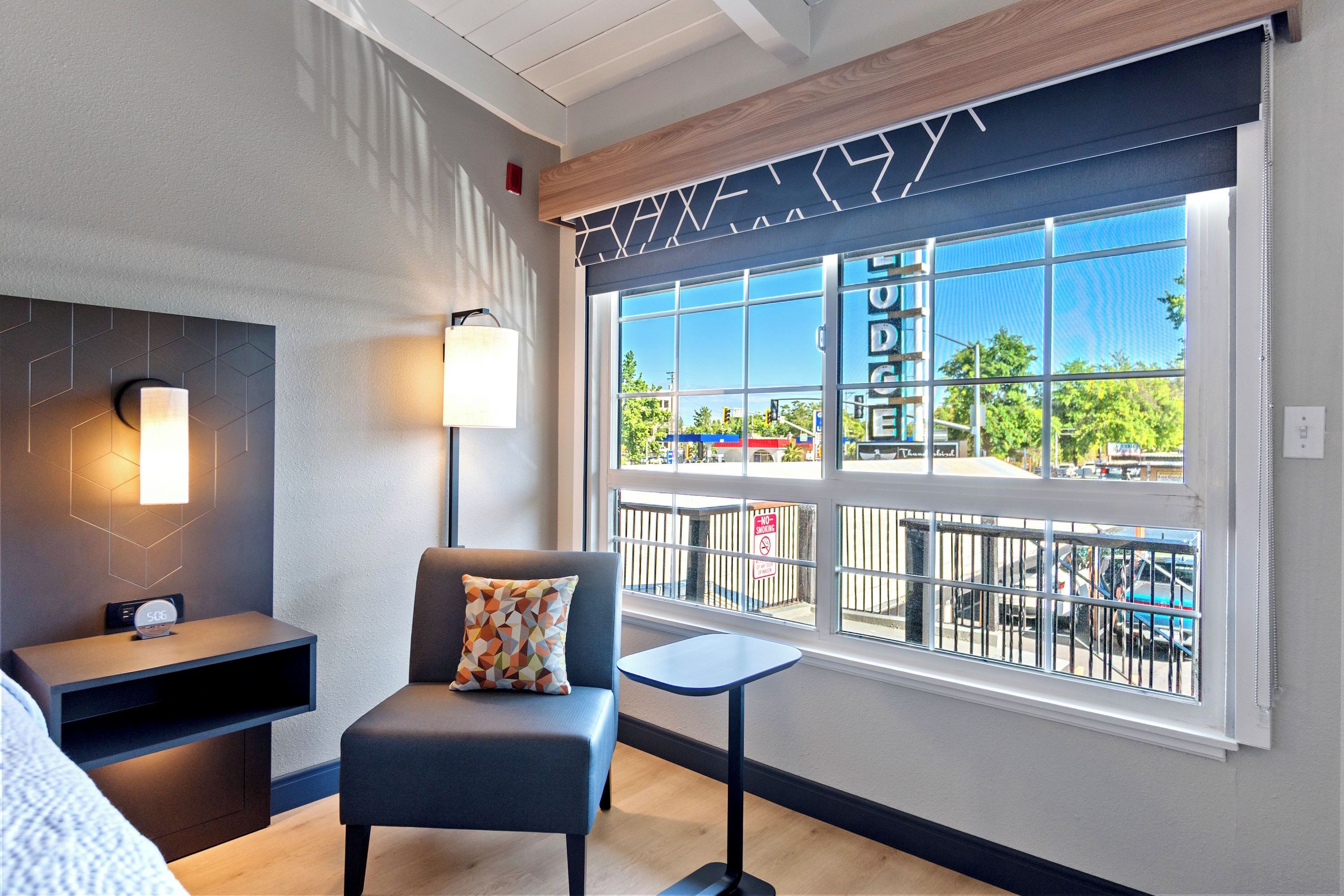 A modern hotel room with a large window, a sofa, a small table, and a wall-mounted lamp.