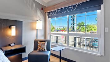 A modern hotel room with a large window, a sofa, a small table, and a wall-mounted lamp.