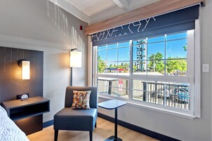 A modern hotel room with a large window, a sofa, a small table, and a wall-mounted lamp.