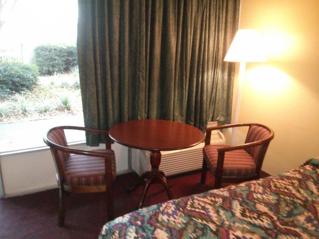 A hotel room with a bed, a round table, two chairs, a lamp, and a view of greenery outside.