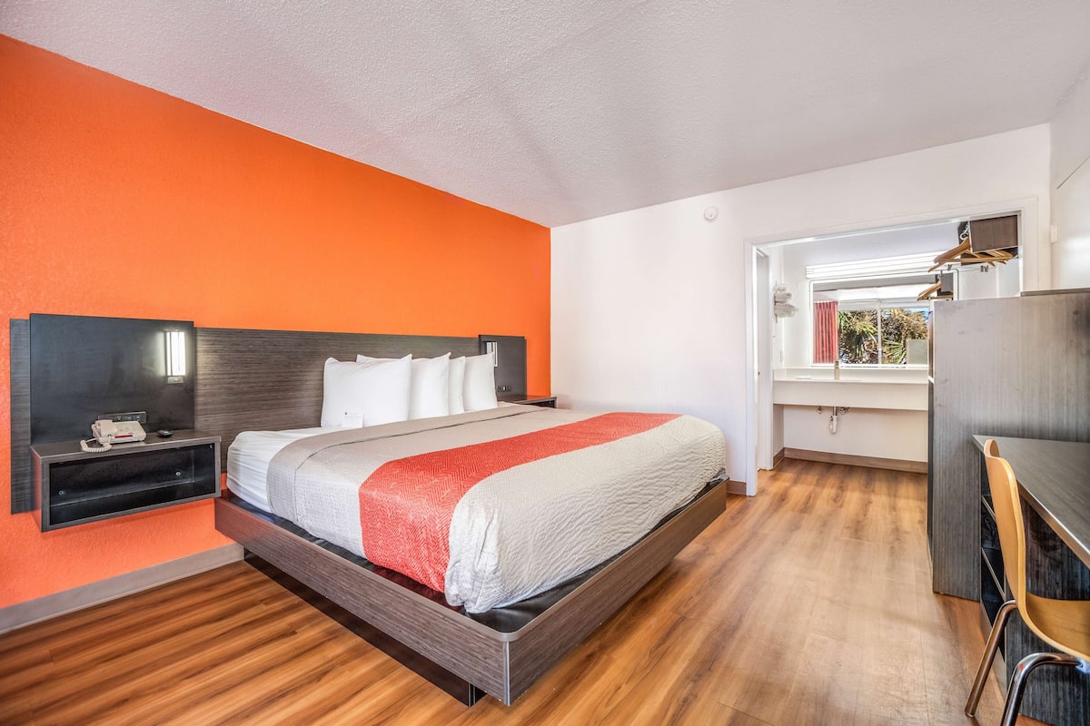 A modern hotel room with a large bed, a bedside table with a lamp, and a view of the outdoors through a window.