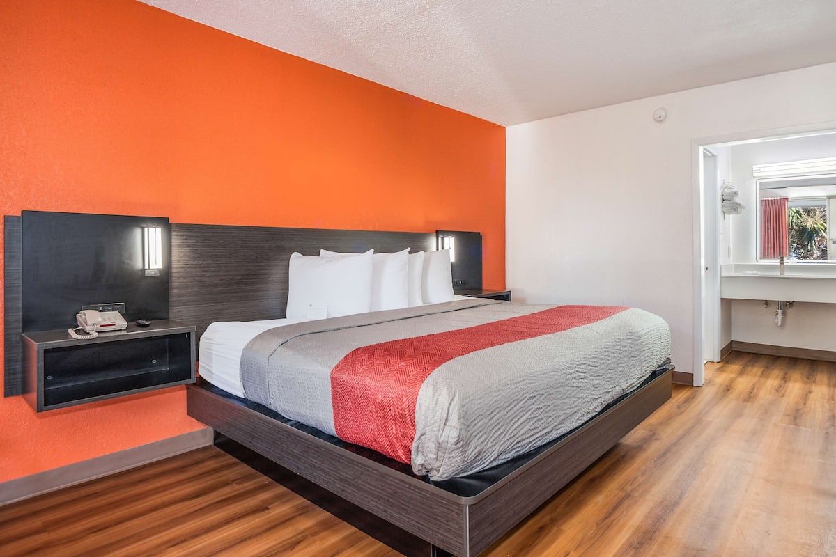 A modern hotel room with a wooden floor, a bed with a grey and red blanket, a nightstand with a phone, and a window with a curtain.