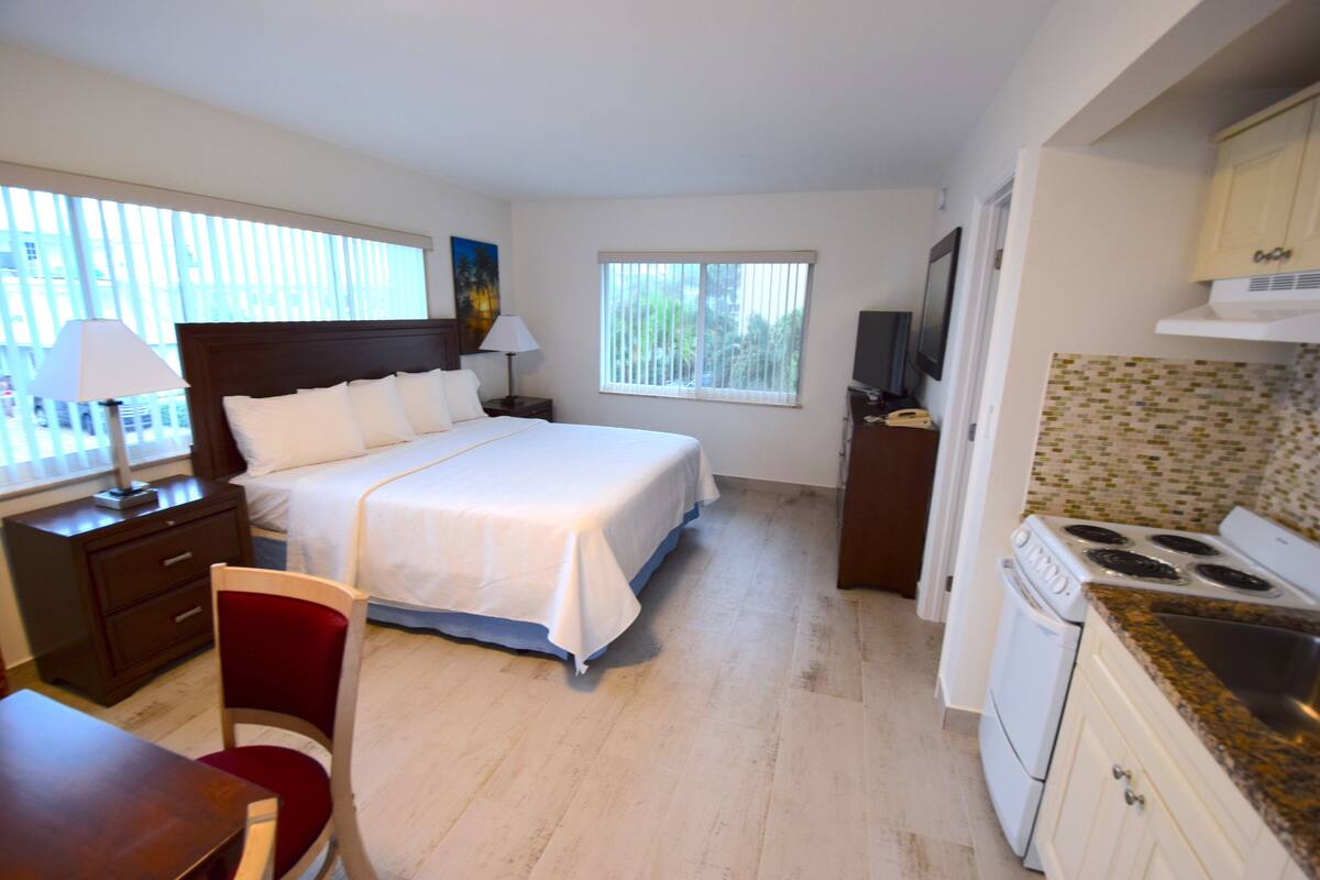 A hotel room with a bed, a desk, a chair, a TV, a kitchen area with a stove and oven, and a window with blinds.