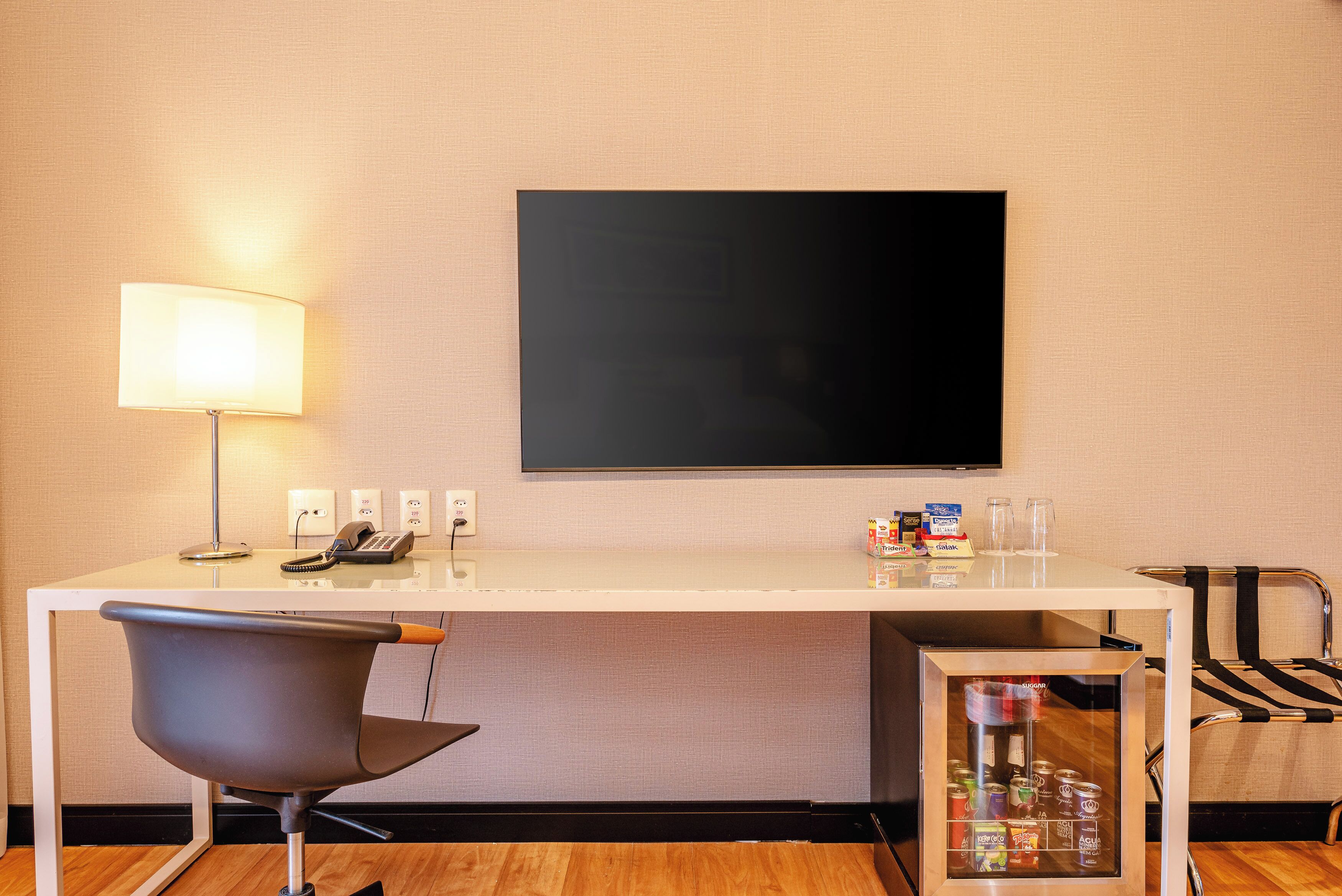 A hotel room with a desk, chair, lamp, flat-screen TV, and a minibar.