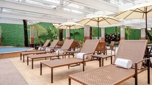 A sunlit indoor pool area with lounge chairs and umbrellas, surrounded by greenery and a glass ceiling. - Blue Tree Premium Verbo Divino (São Paulo)