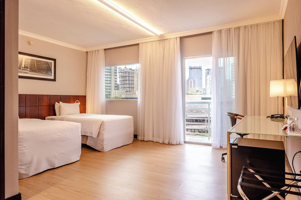Une chambre d’hôtel avec deux lits, un bureau avec une chaise, une grande fenêtre avec des rideaux et un tableau encadré au mur.
