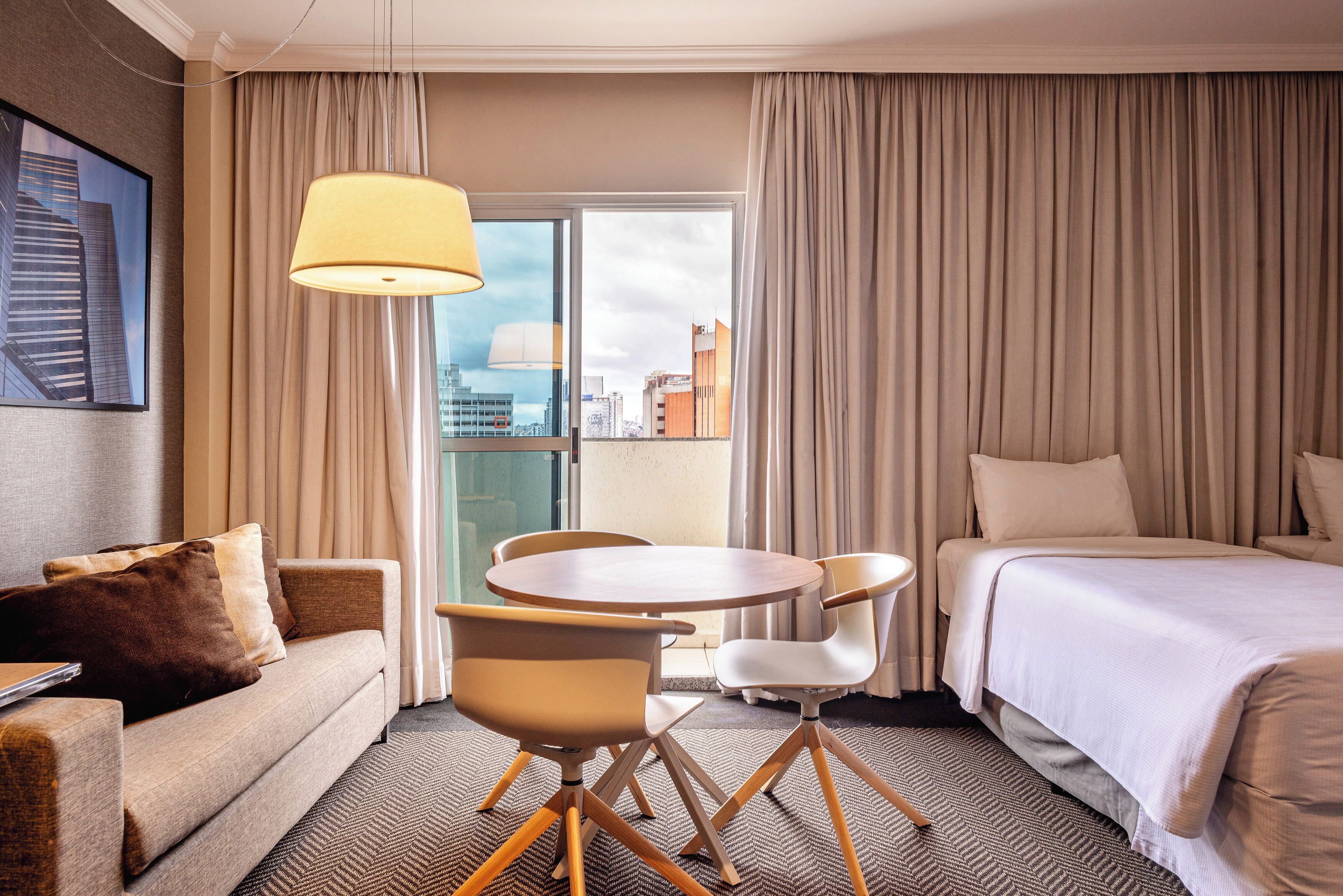 A hotel room with a bed, a sofa, a dining table with chairs, a lamp, and a view of a cityscape through a large window.