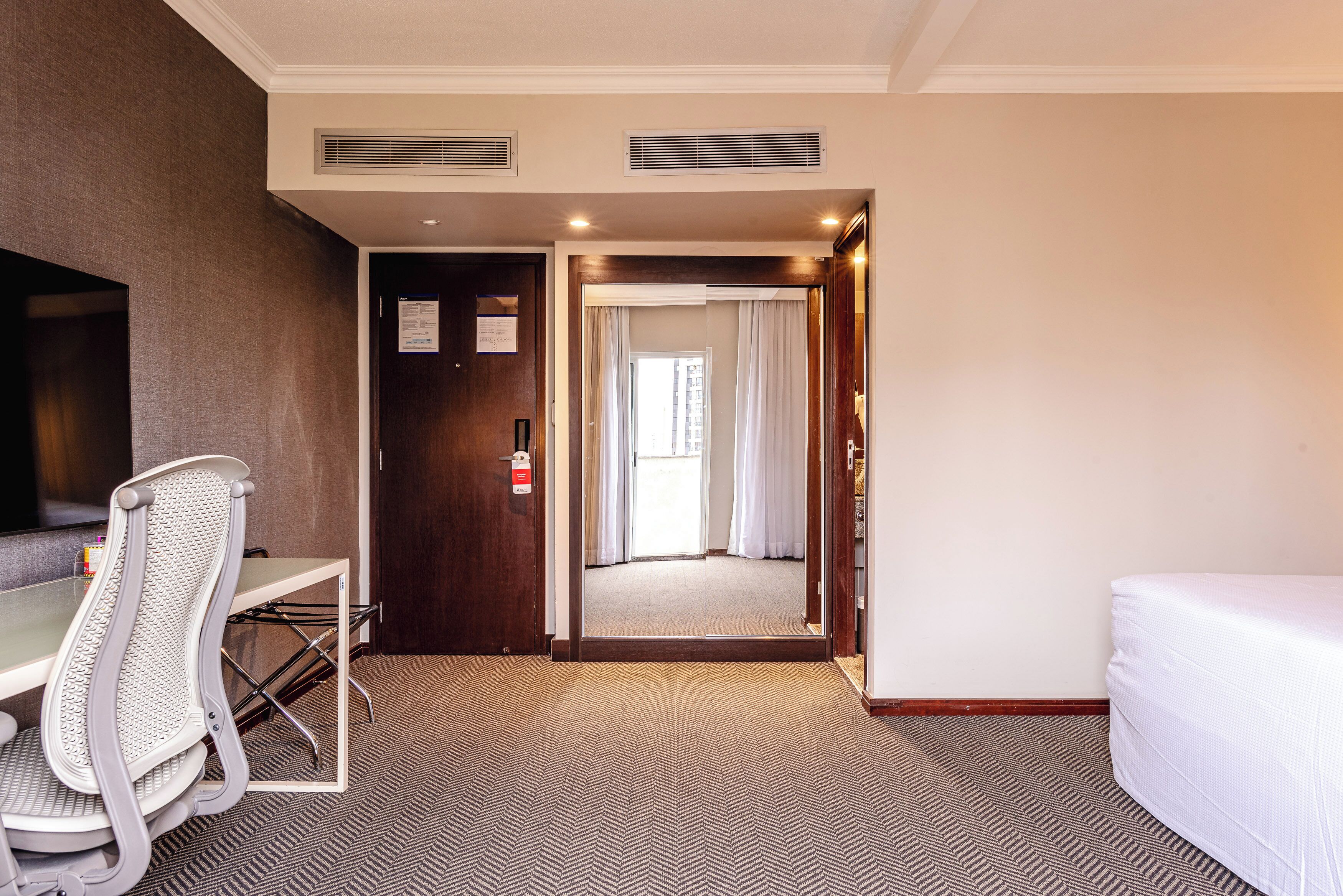 A hotel room with a bed, a desk, and a chair. There is a door leading to a hallway with a mirror and a window.