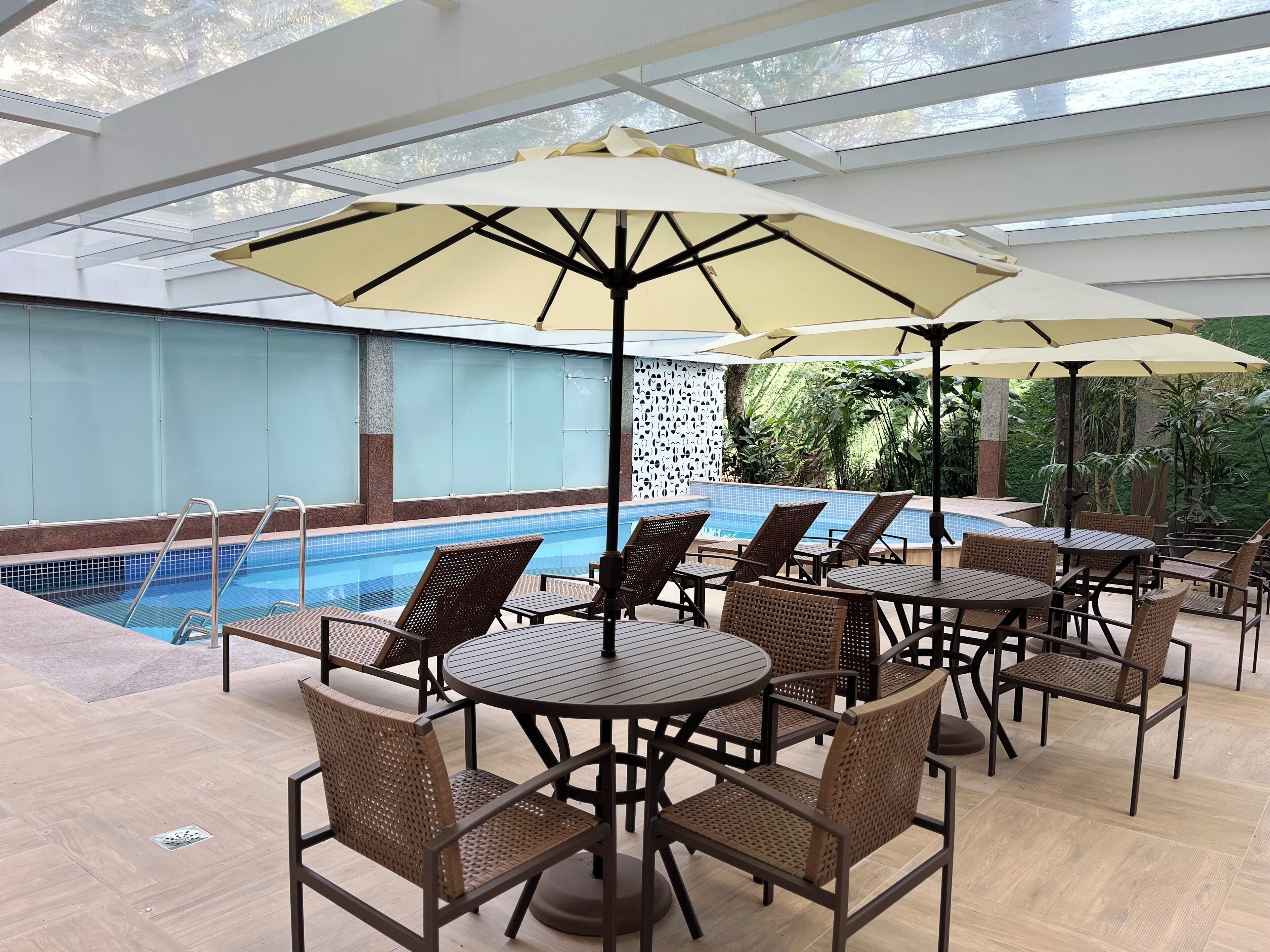 A poolside area with lounge chairs and tables, a swimming pool, and a glass ceiling.