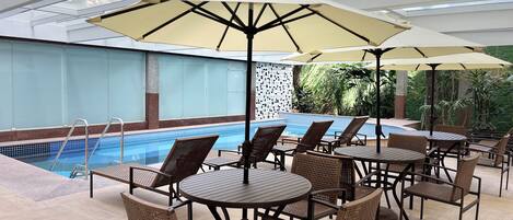 A poolside area with lounge chairs and tables, a swimming pool, and a glass ceiling.