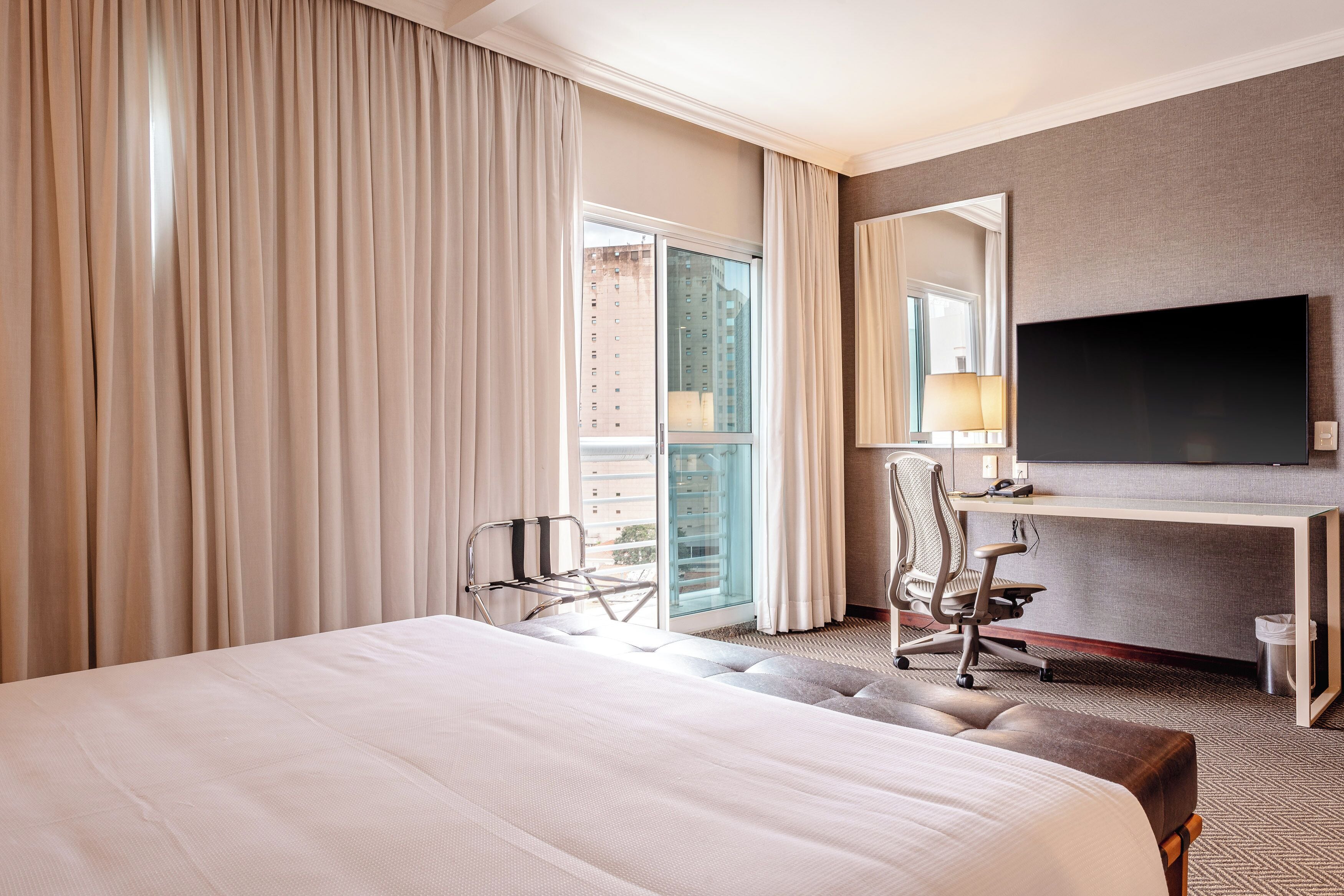 A hotel room with a large bed, a desk with a chair, a television, and a view of a building through the window.