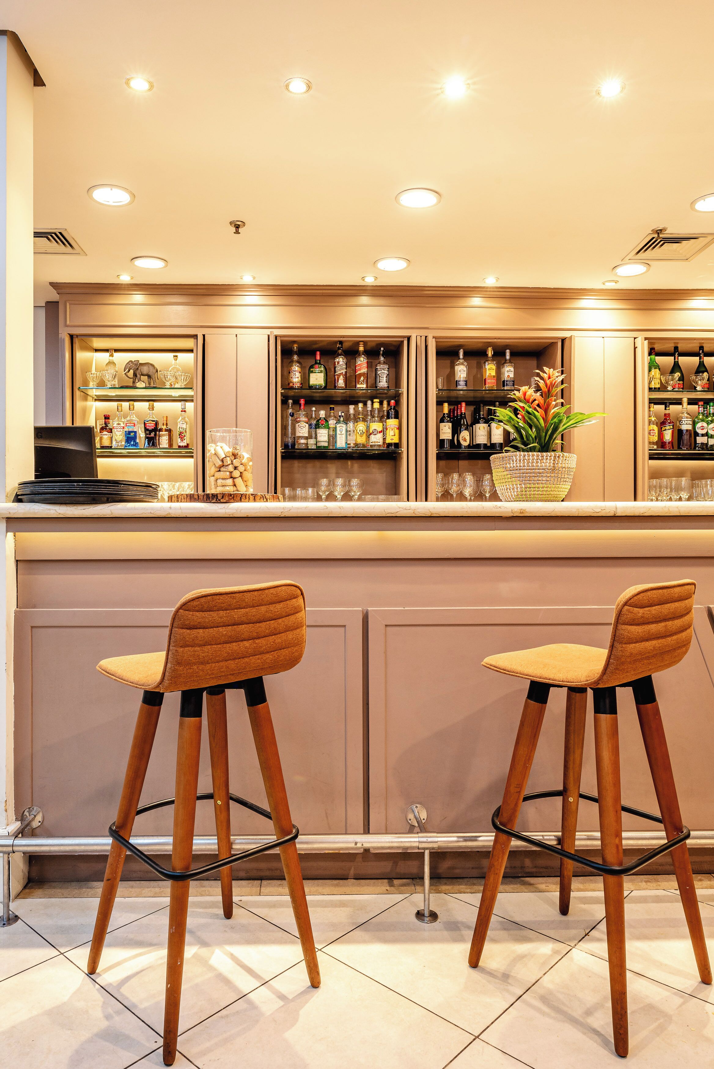 A bar area with two wooden bar stools, a countertop with a laptop, and shelves stocked with bottles and glasses.