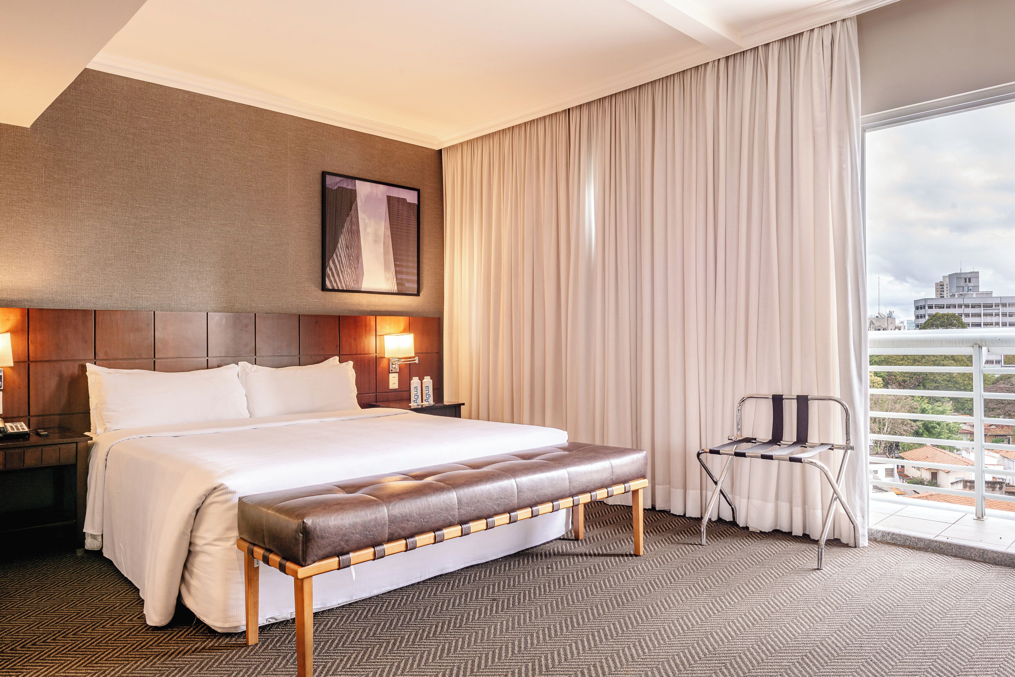 A hotel room with a large bed, a bench, a chair, a nightstand, and a view of the city through the window.