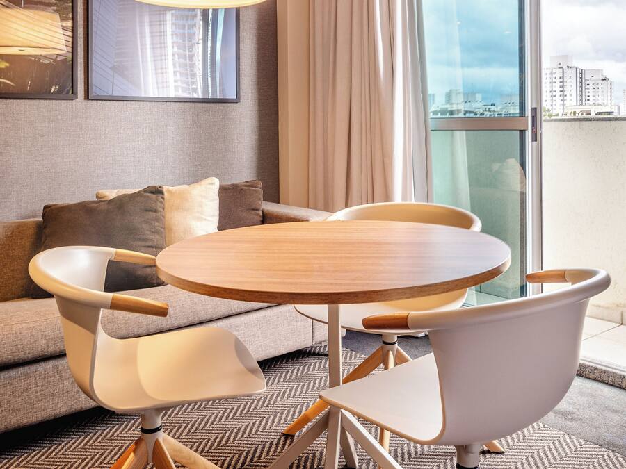 Ein modernes Hotelzimmer mit einem runden Holztisch, weißen Stühlen, einer Couch und einem großen Fenster mit Blick auf die Stadt.