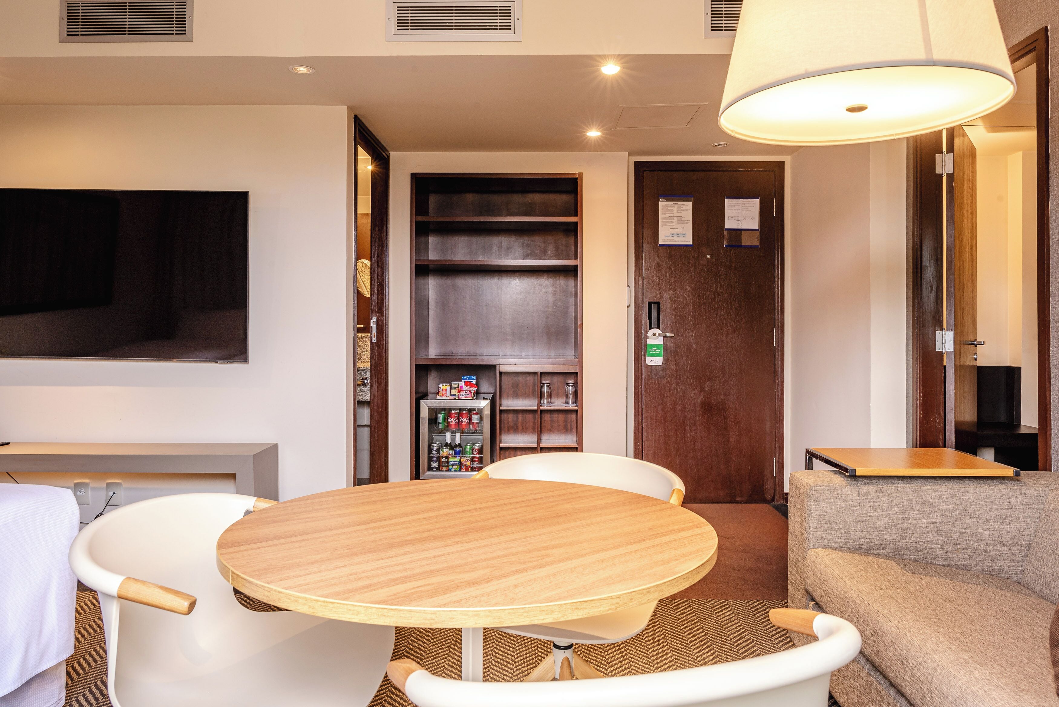 Habitación de hotel con una mesa redonda de madera, sillas blancas, un sofá, un televisor, una estantería y una puerta con un cartel.
