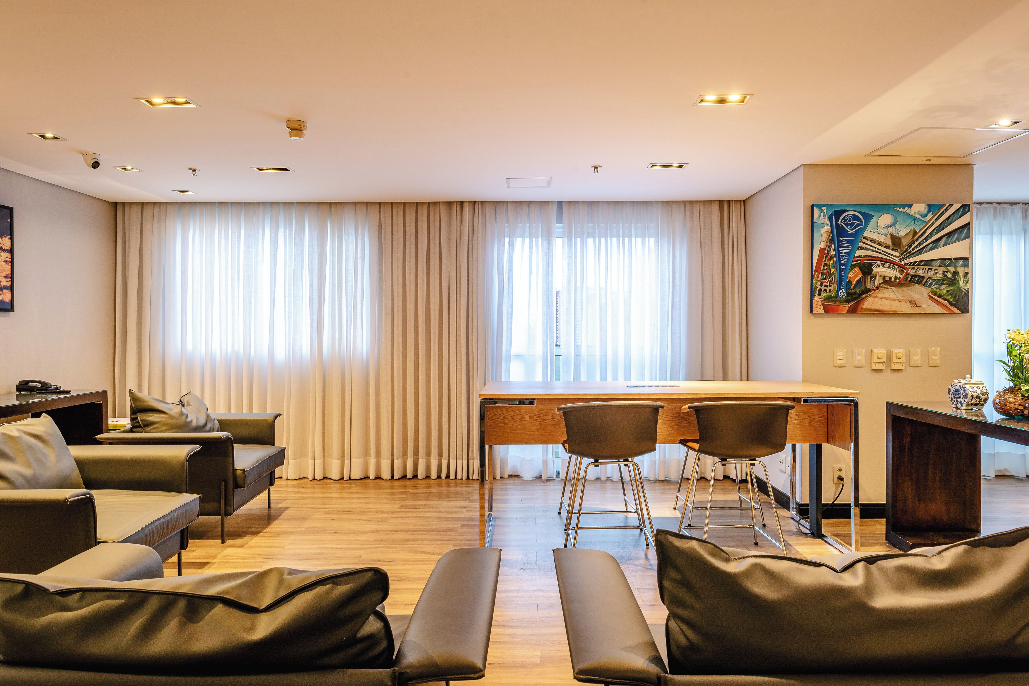 A modern hotel room with a wooden desk, two chairs, a sofa, and a large window with curtains.