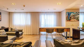 A modern hotel room with a wooden desk, two chairs, a sofa, and a large window with curtains.
