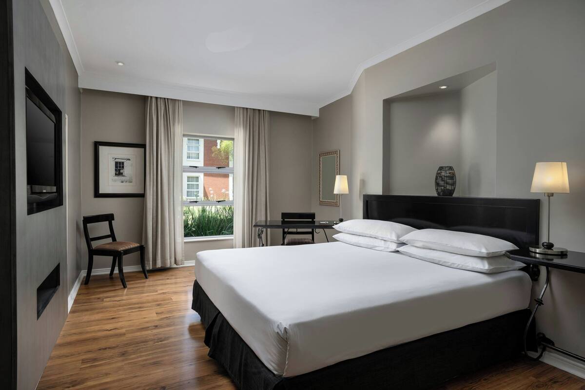 A modern hotel room with a large bed, a desk, a chair, and a window with curtains.