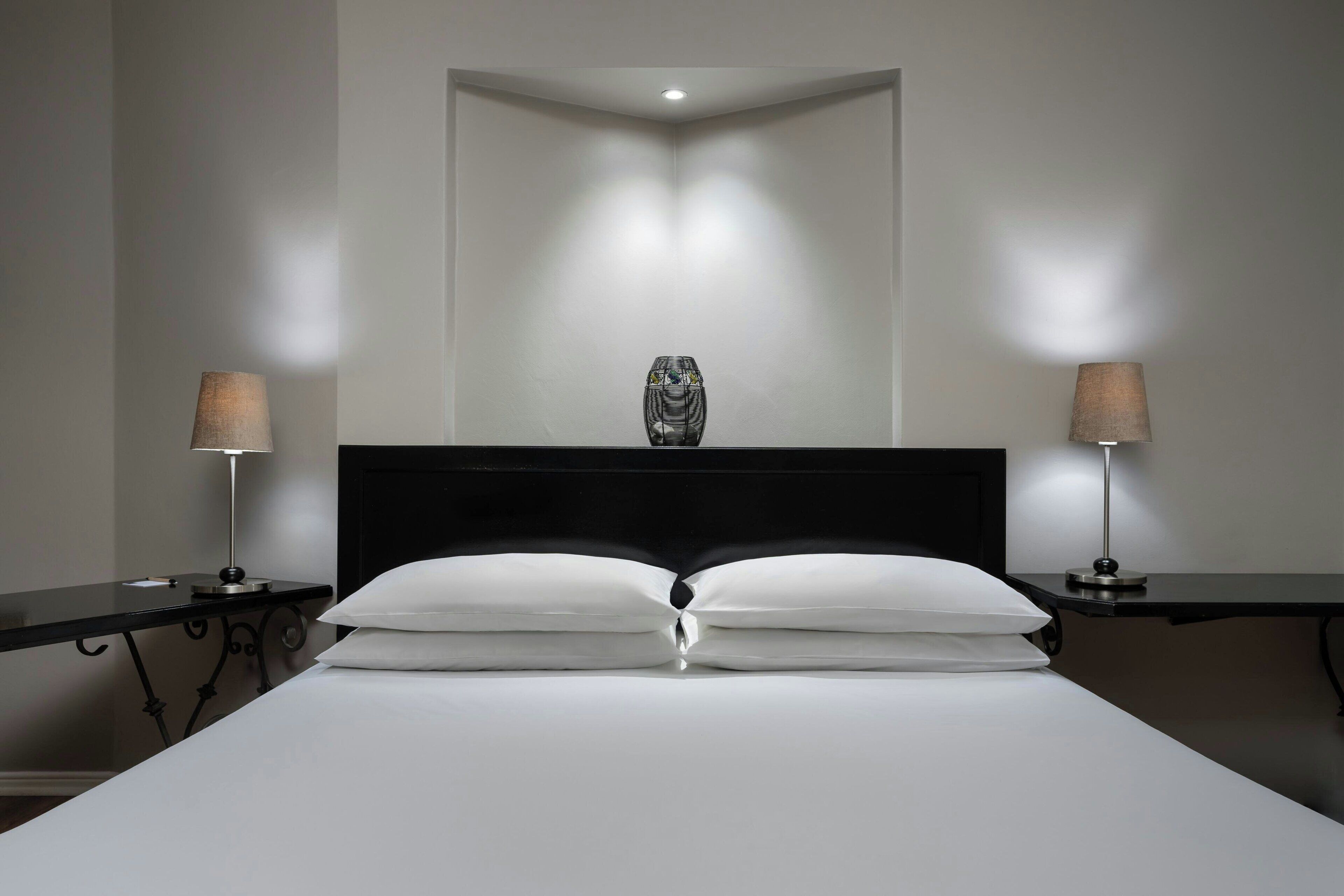 Una cama bien tendida con sábanas blancas y dos almohadas, flanqueada por dos mesitas de noche con lámparas, en una habitación con cabecera oscura y una sola lámpara en el techo.