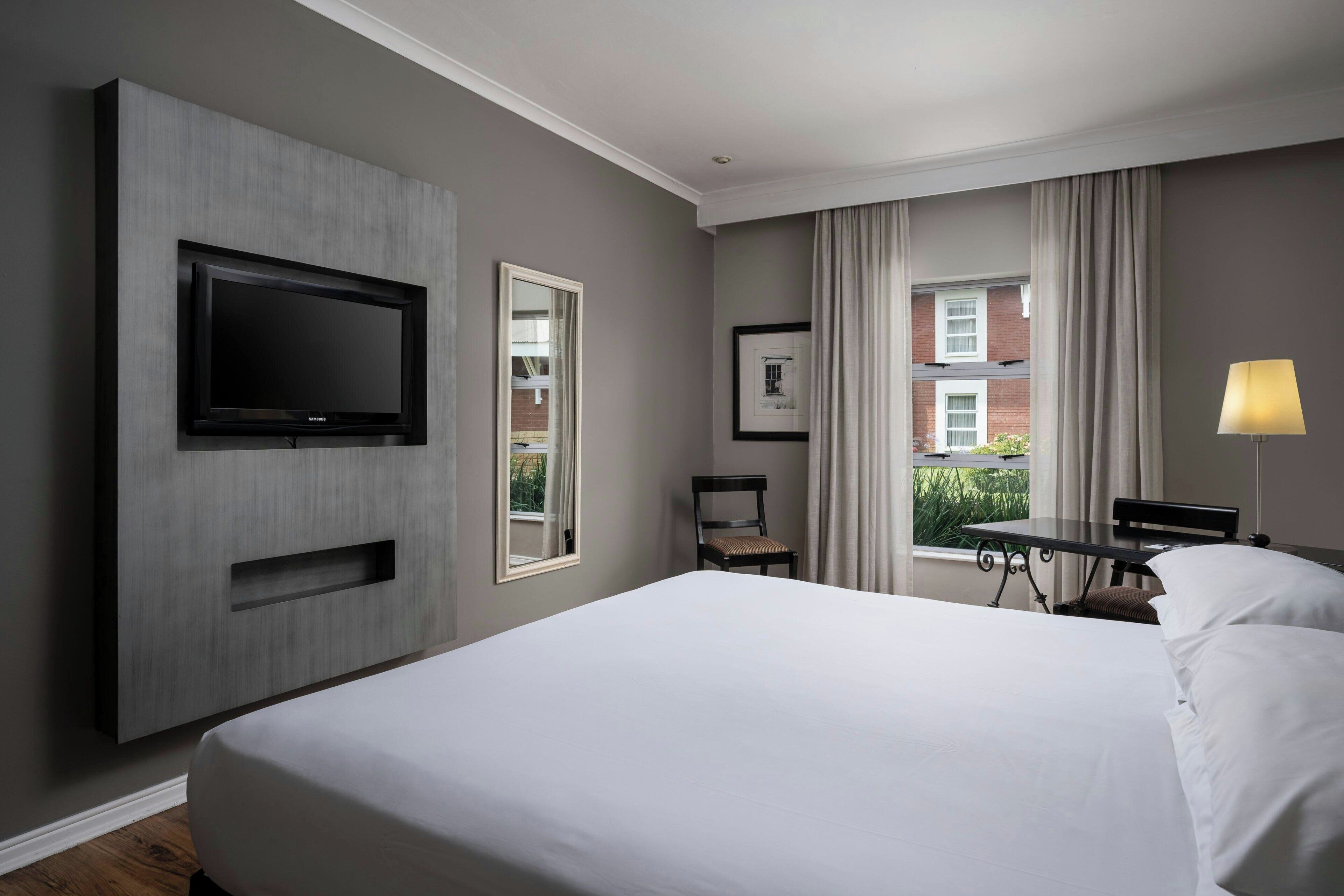 Una habitación de hotel con una cama grande, un televisor de pantalla plana montado en la pared, una mesita con una silla y una ventana con cortinas.