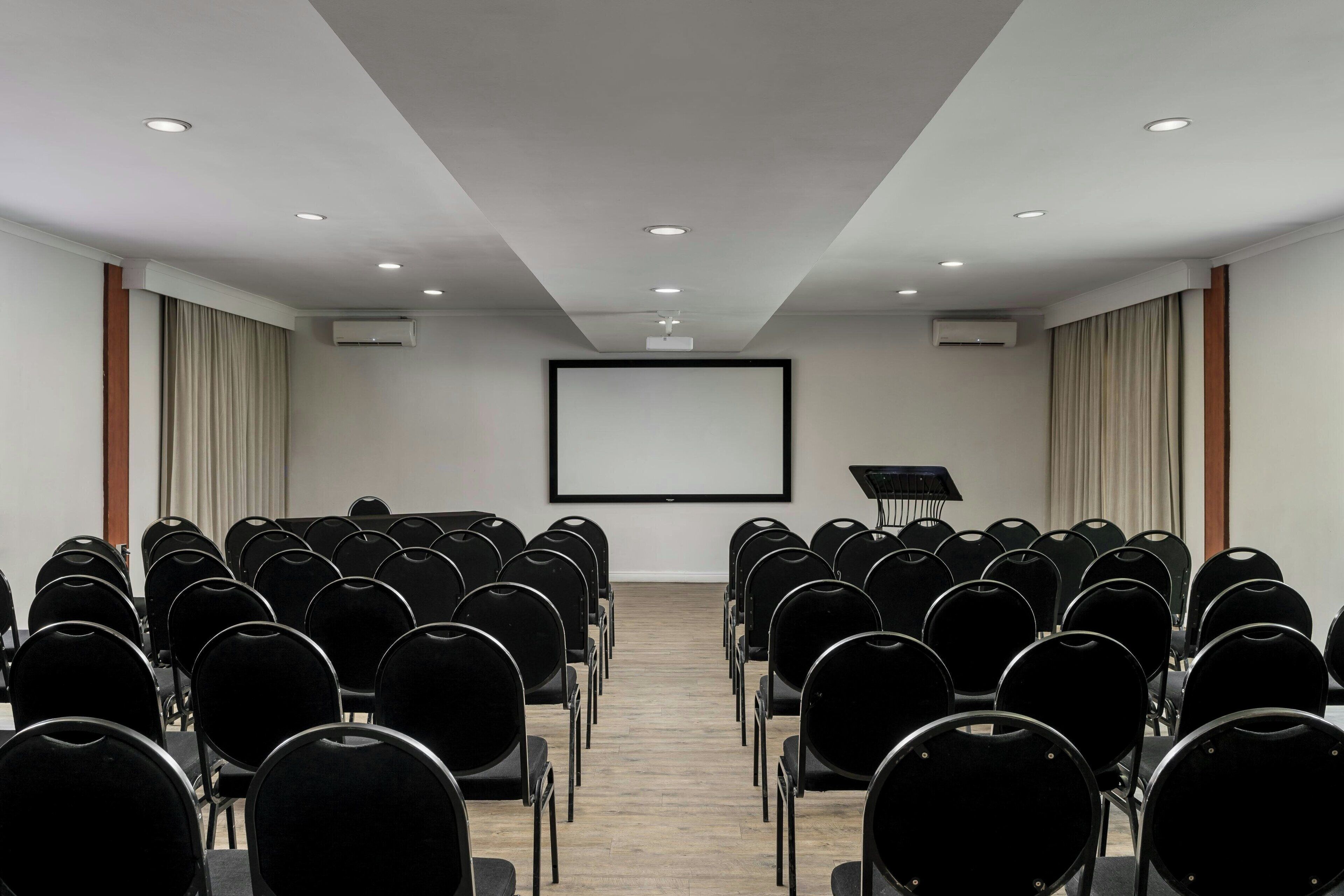 Une salle de conférence avec des rangées de chaises noires disposées face à un grand écran blanc, un plancher en bois et des luminaires au plafond.