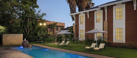 A swimming pool with lounge chairs and umbrellas, surrounded by a brick building and palm trees.