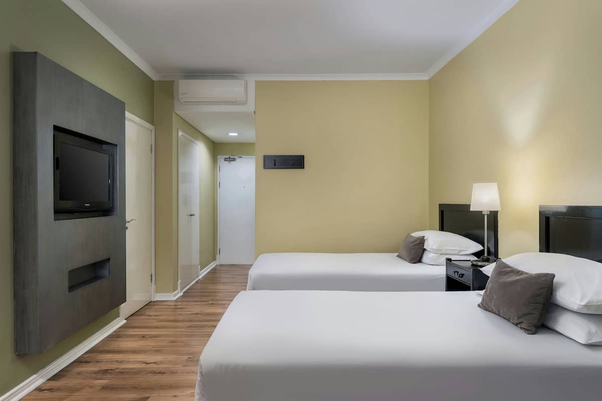 A hotel room with two beds, a TV mounted on the wall, a bedside table with a lamp, and a door leading to another room.