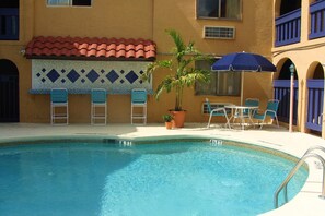 Une piscine avec un bord carrelé, un palmier et un espace extérieur aménagé avec des parasols.