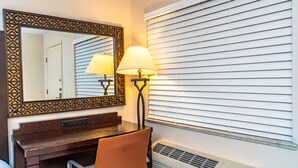 A hotel room with a wooden desk, two lamps, a mirror, and a window with blinds. - Sevilla Inn - Near Medieval Times (Kissimmee)