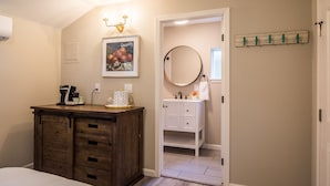 Une chambre avec une commode en bois, un lit, une table de chevet, un tableau encadré, un miroir rond et une salle de bain visible à travers une porte ouverte.