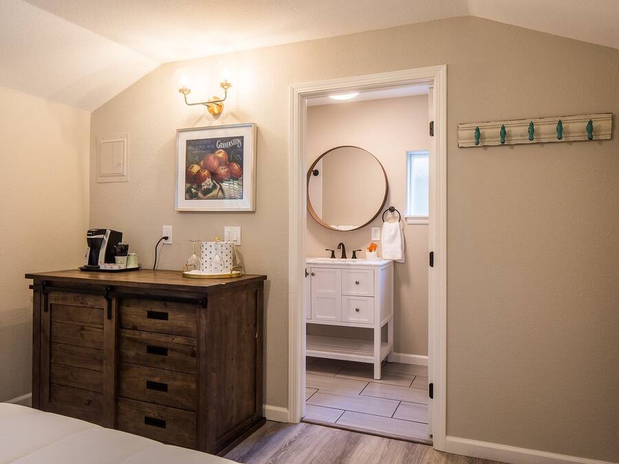 Une chambre avec une commode en bois, un lit, une table de chevet, un tableau encadré, un miroir rond et une salle de bain visible à travers une porte ouverte.