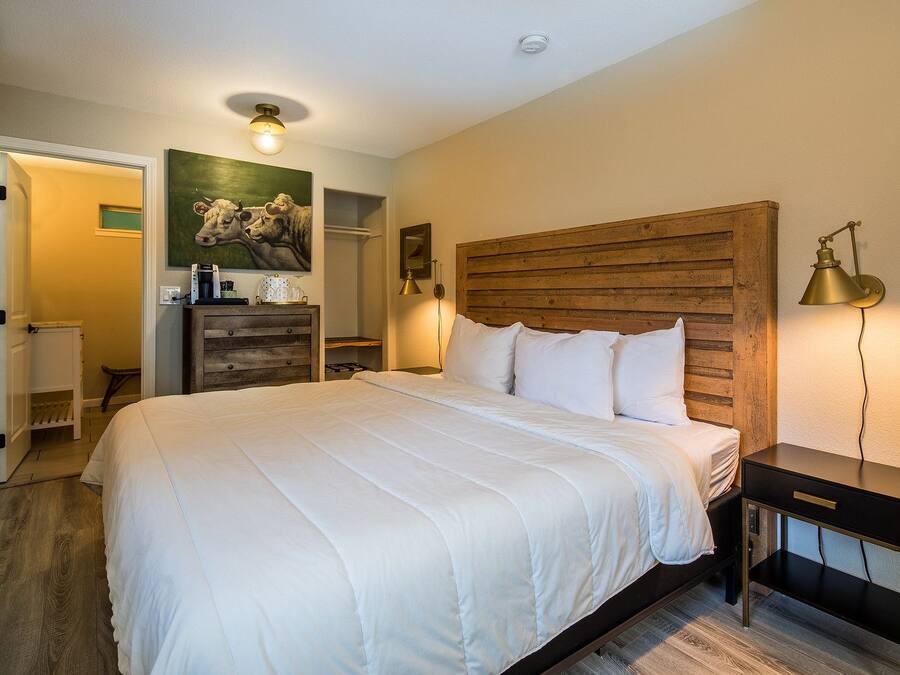 A bedroom with a large bed, a wooden headboard, a nightstand, a lamp, a painting on the wall, and a door leading to another room.