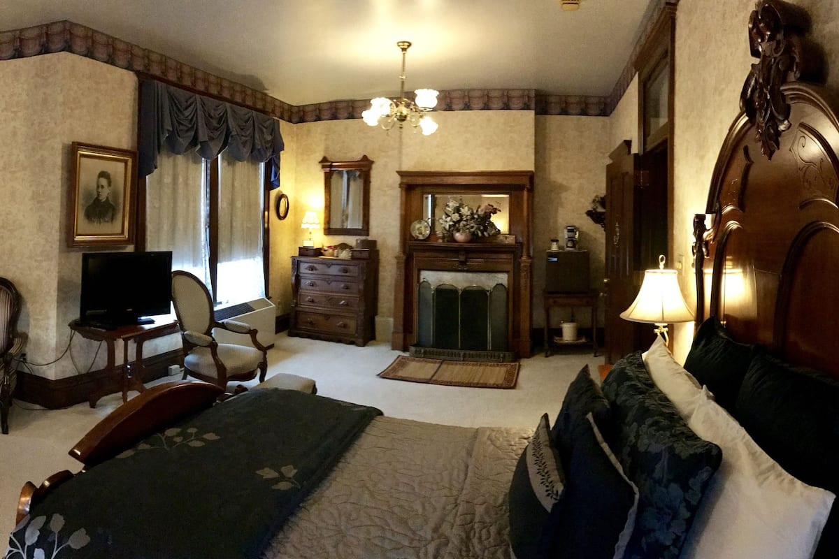 A traditional hotel room with a sofa, armchair, bed, television, and a fireplace.