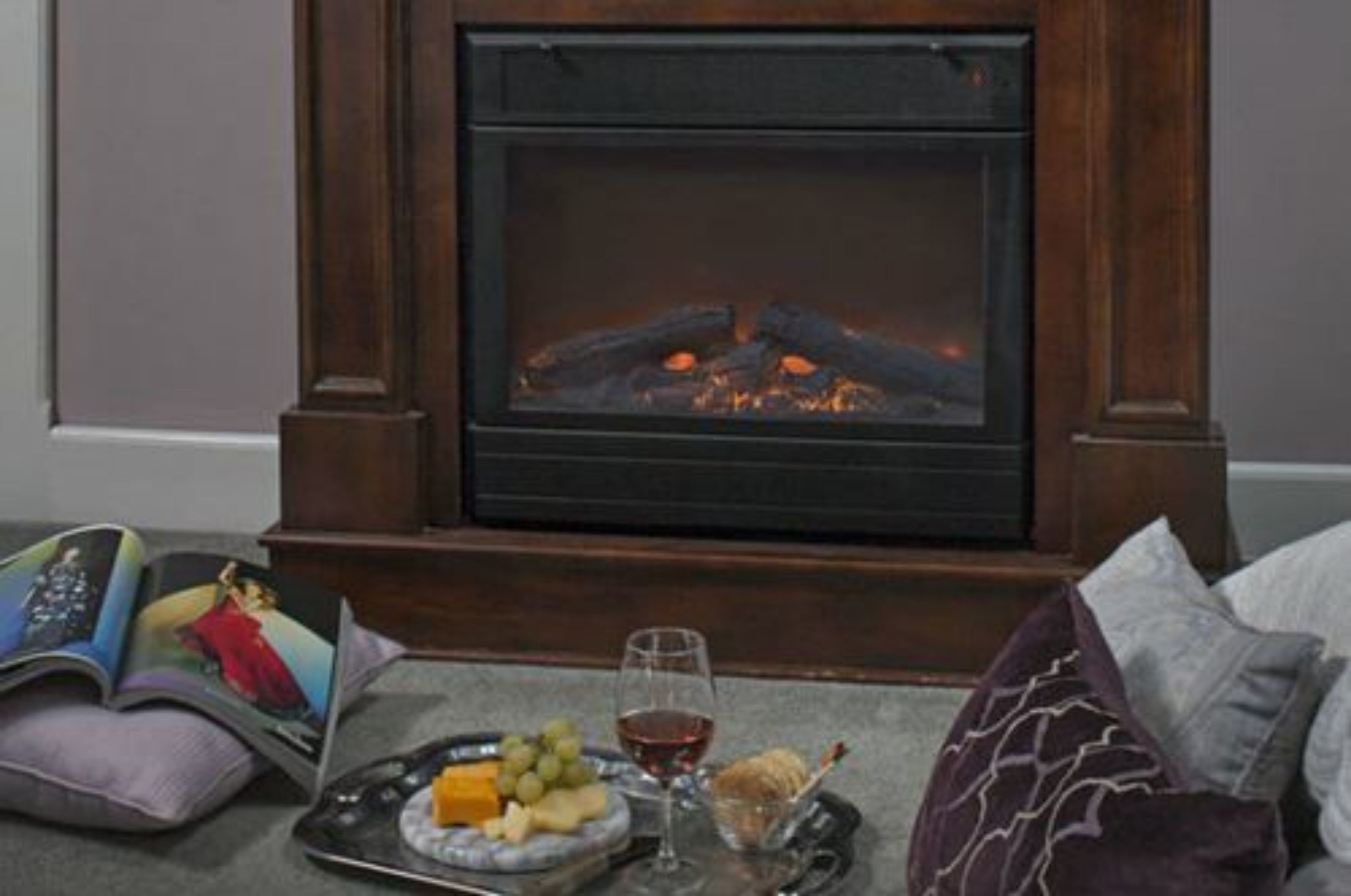 Una chimenea con fuego, una bandeja con queso, uvas y una copa de vino, y un libro sobre una mesa.