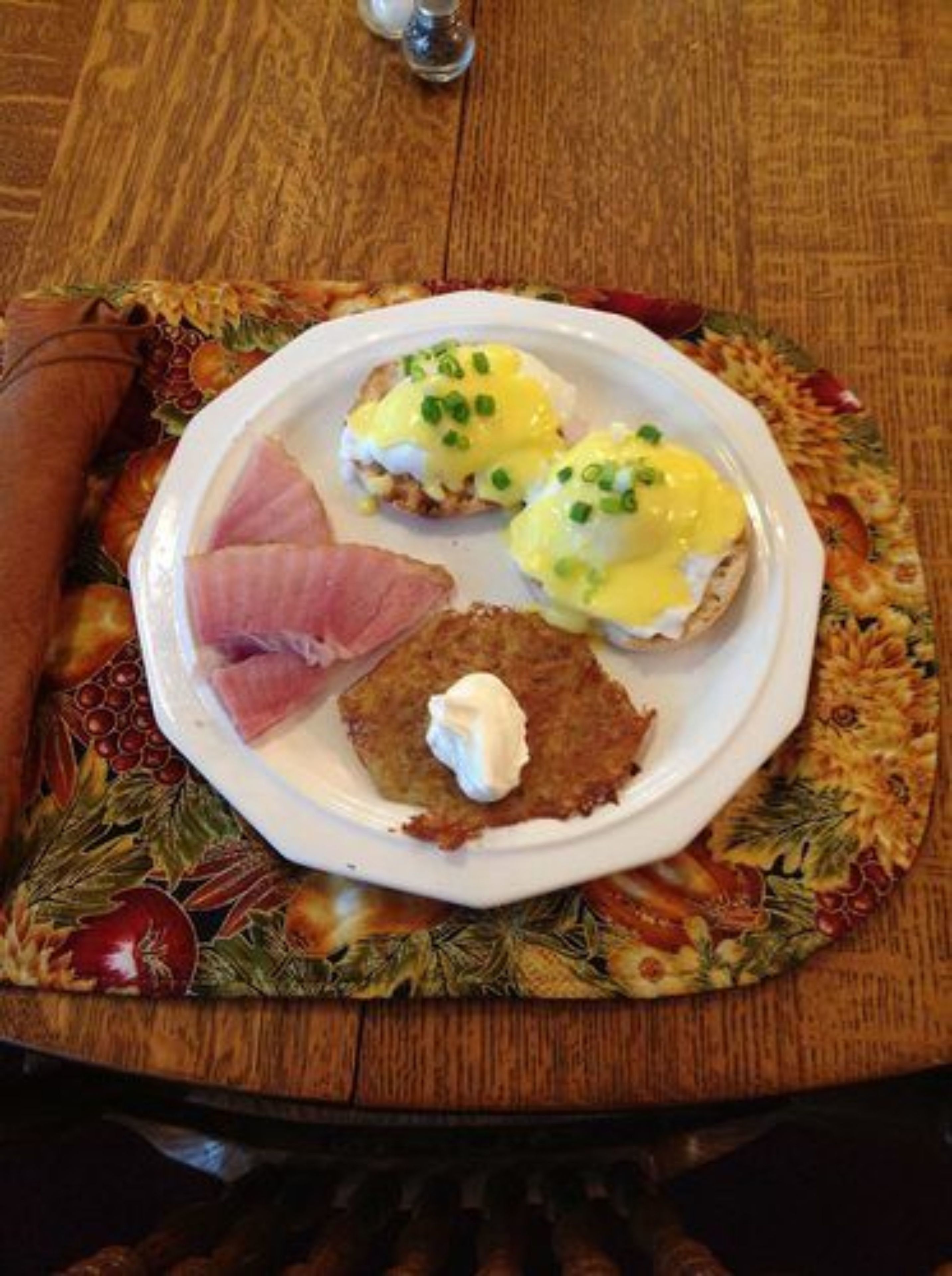 Une assiette contenant des œufs Bénédicte et une galette de pommes de terre, posée sur une table recouverte d’un set de table à motifs floraux.