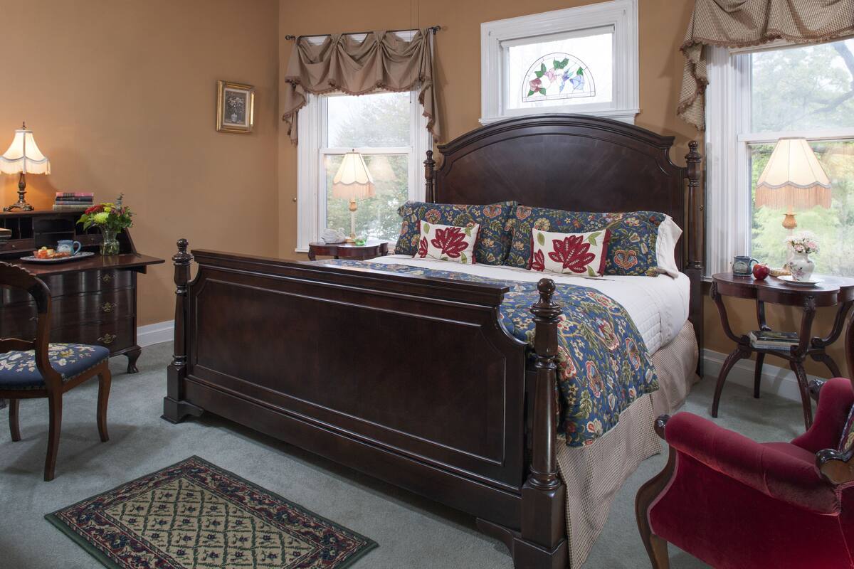 A bedroom with a large bed, a chair, a desk, and a window with curtains.