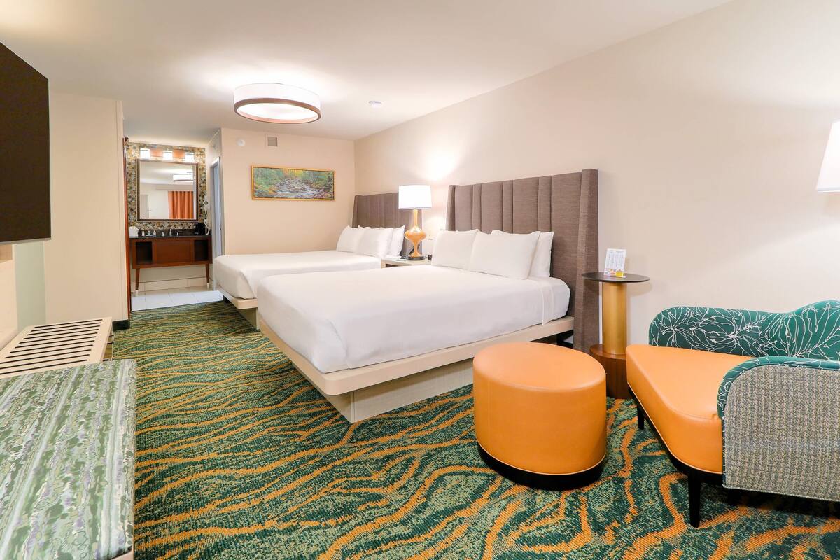 A hotel room with a large bed, two bedside tables, a sofa, an ottoman, a mirror, and a bathroom visible in the background.