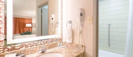 A bathroom with a granite countertop sink, a large mirror, a hairdryer, and a shower.