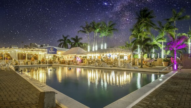 A nighttime pool area with a clear view of the pool, surrounding palm trees, and a building with a sign.