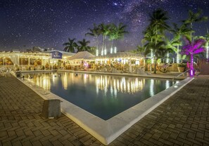 Zona de piscina nocturna con vista clara al agua, palmeras circundantes y un edificio con un letrero.