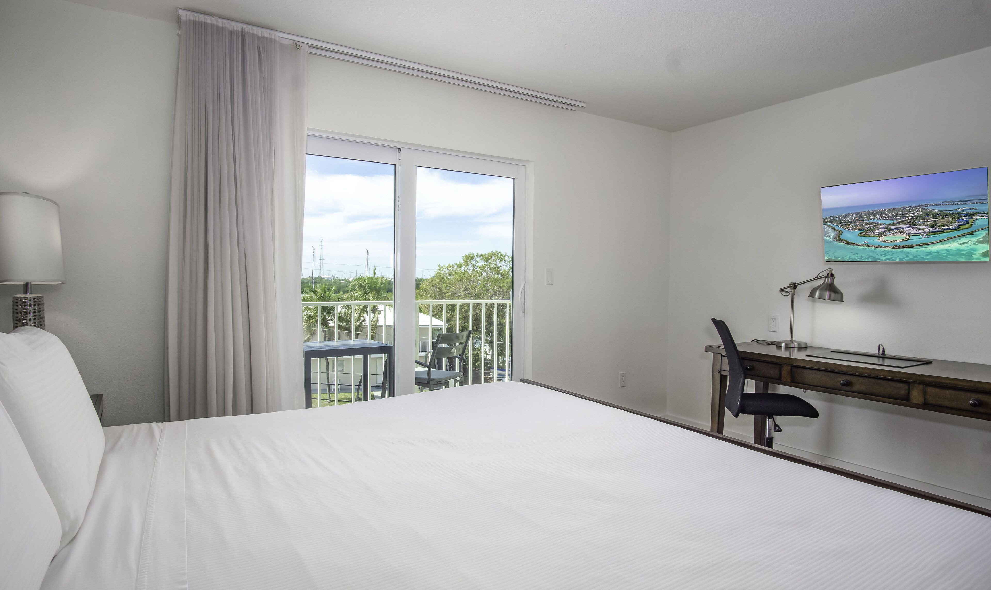 A hotel room with a large bed, a desk with a chair, a TV on the wall, and a balcony with a view of the sea.