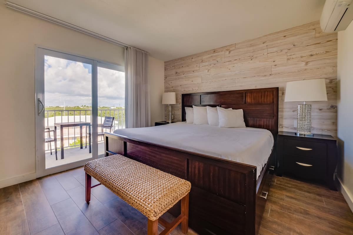 Ein Hotelzimmer mit einem großen Bett, einem hölzernen Kopfteil, einem Nachttisch mit Lampe, einer Bank und einem Balkon mit Blick auf den Himmel.