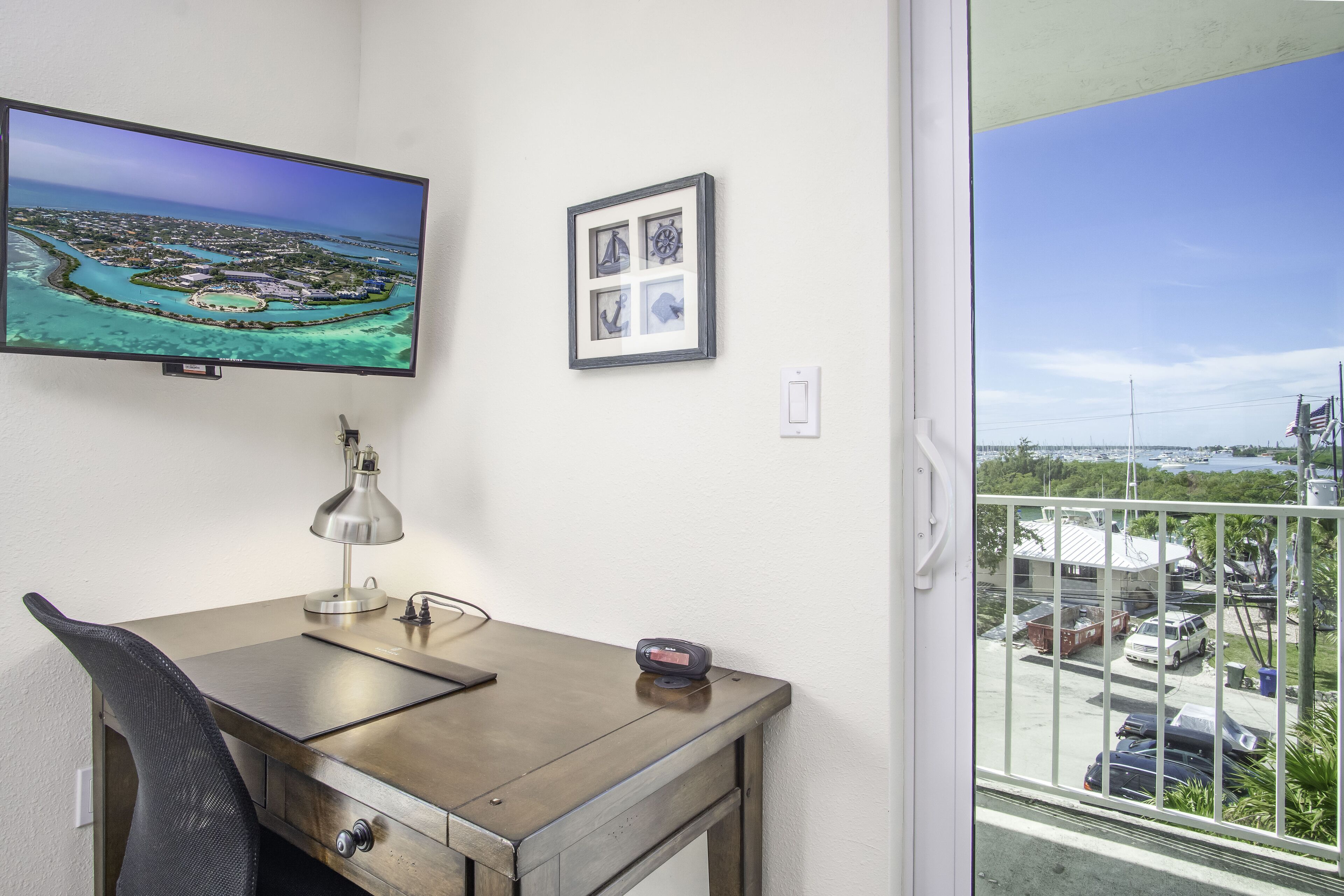 A room with a desk, a chair, a television, a framed picture, and a window with a view of a marina.
